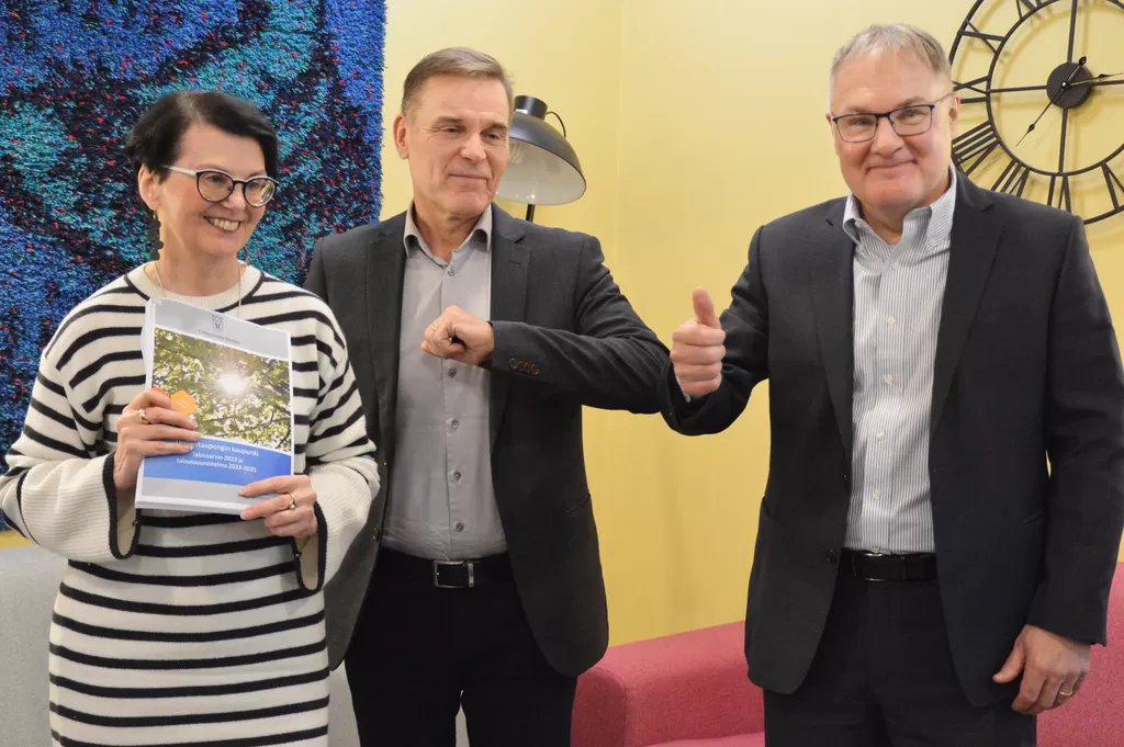Anne Takala, Raimo Löfstedt ja Atso Vainio esittelevät ensi vuoden budjettikirjaa. Kanteen on valittu aurinkoinen kuva.